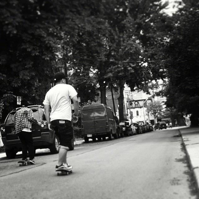 People skateboarding on a city street with parked cars and trees lining the sidewalk.