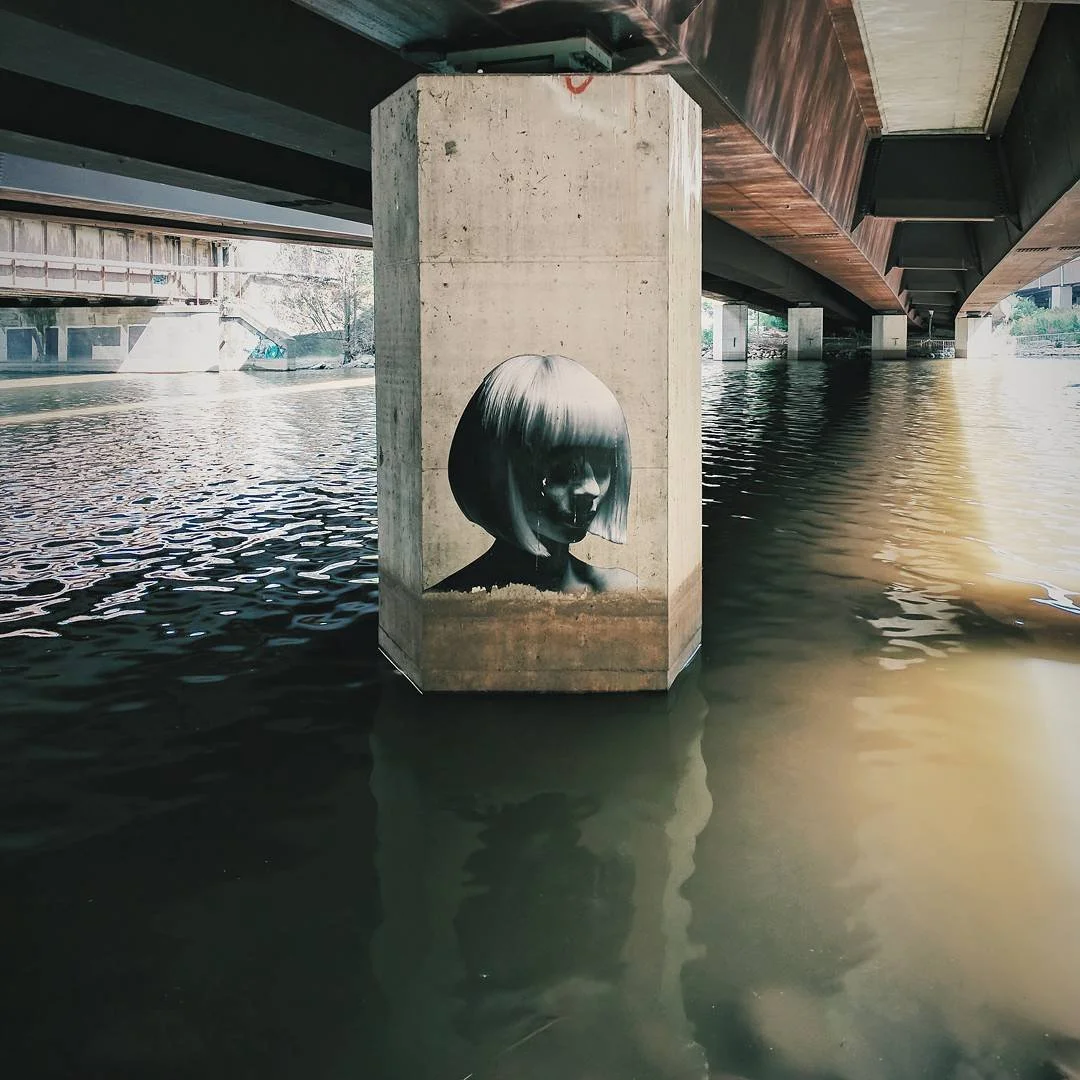 Street art graffiti of a black and white portrait of a woman with a bob haircut painted on a concrete pillar under a bridge, partially submerged in water.