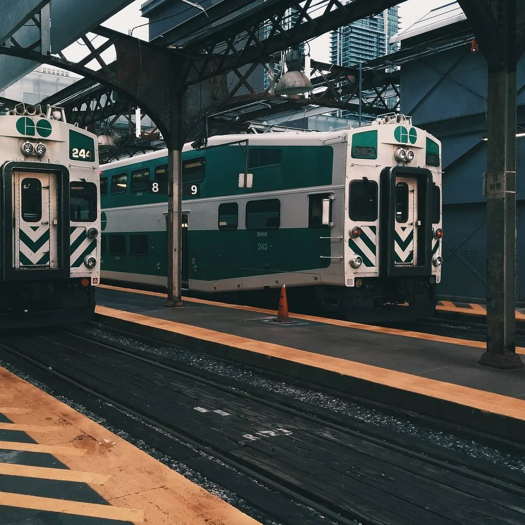 Two electric trains parked at a train station platform with overpass structure above, surrounded by modern tall buildings.