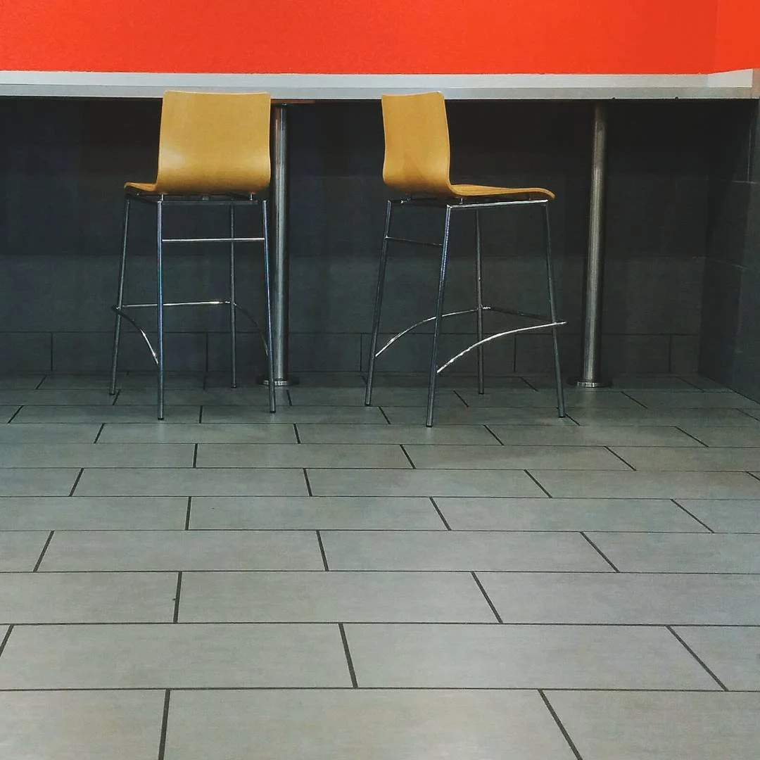 Two yellow bar stools in front of a high table in an indoor setting, with gray tiled floor and orange wall in the background.