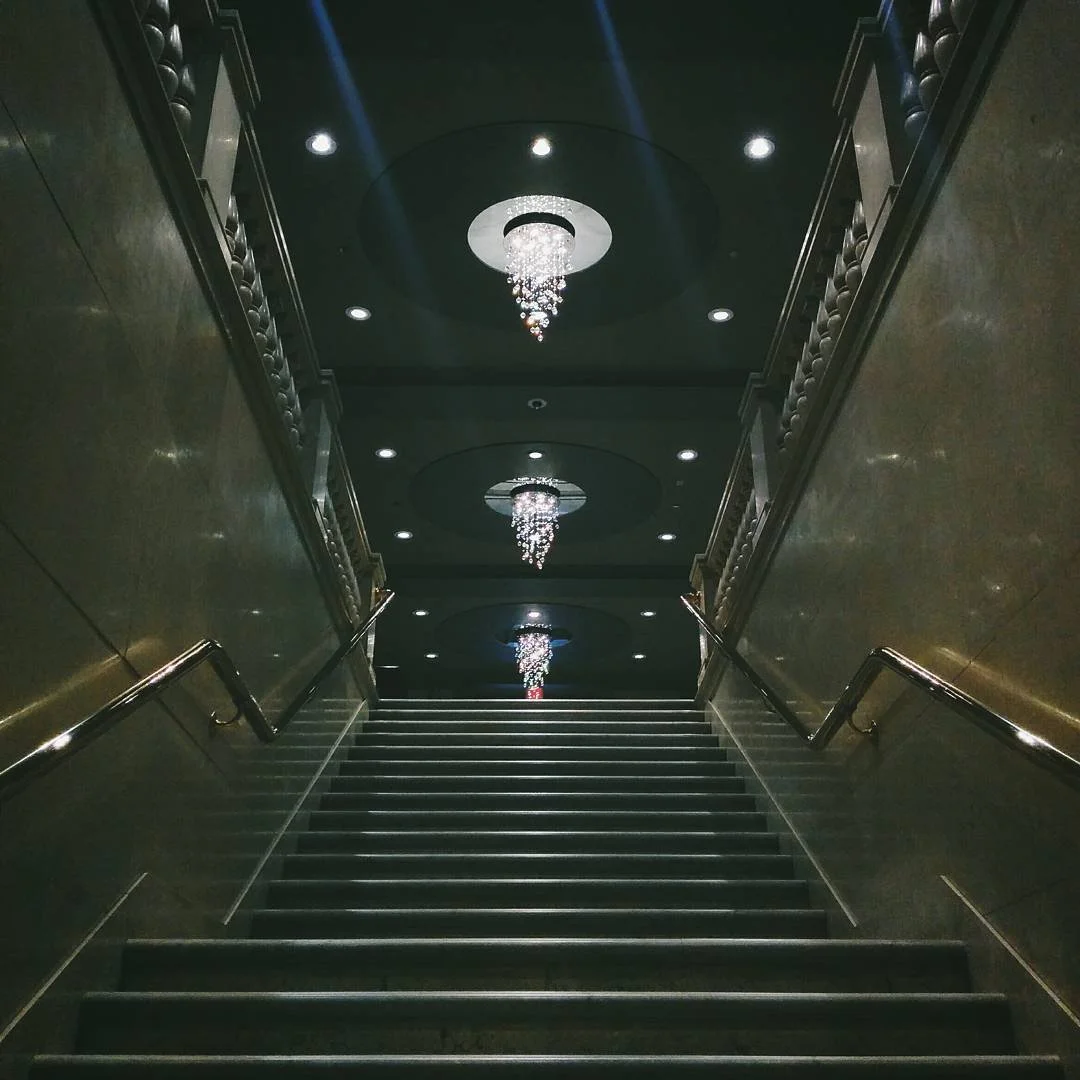 Elegant interior staircase leading upward, with polished metal handrails on both sides, dark tiled steps, and a black ceiling featuring three crystal chandeliers aligned along the staircase's center axis.