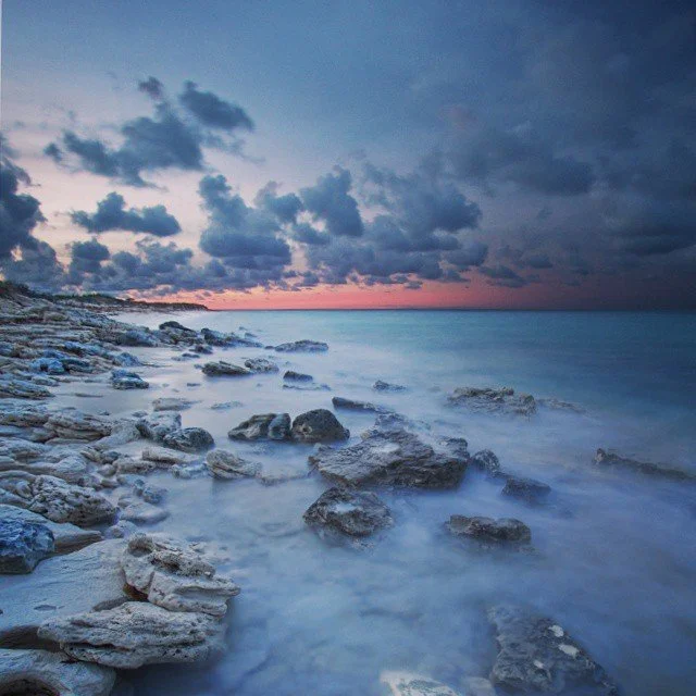 A rocky shoreline during sunset with a pink and purple sky and dark clouds over the ocean.