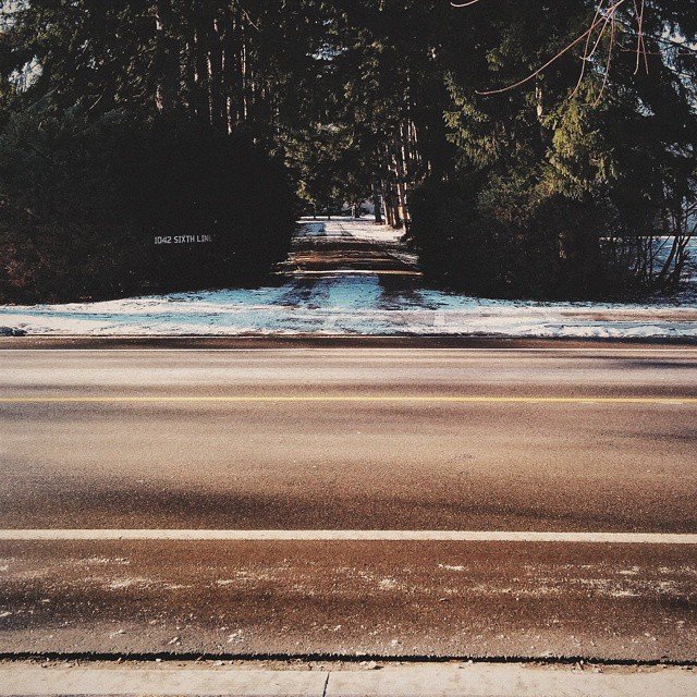 A road with painted lane lines leading to a dirt path surrounded by trees, with snow on the ground and a sign that reads '1042 SIXTH LINK'.