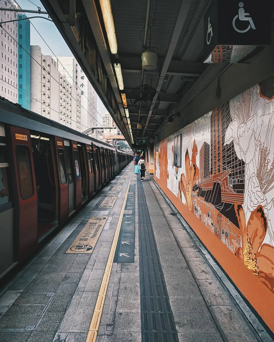 Subway train at an outdoor station in an urban setting. Passengers wait on the platform, which has tiled flooring, tactile paving, and wall art depicting cityscapes and nature. Signage indicates accessibility for disabled individuals.