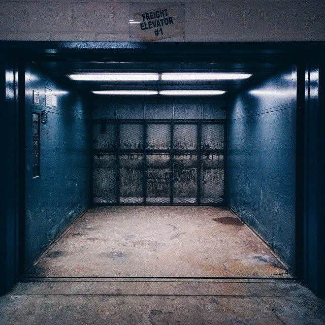 Empty freight elevator with a metal mesh gate at the back, blue walls, fluorescent lighting, and a sign overhead reading 'FREIGHT ELEVATOR #1'