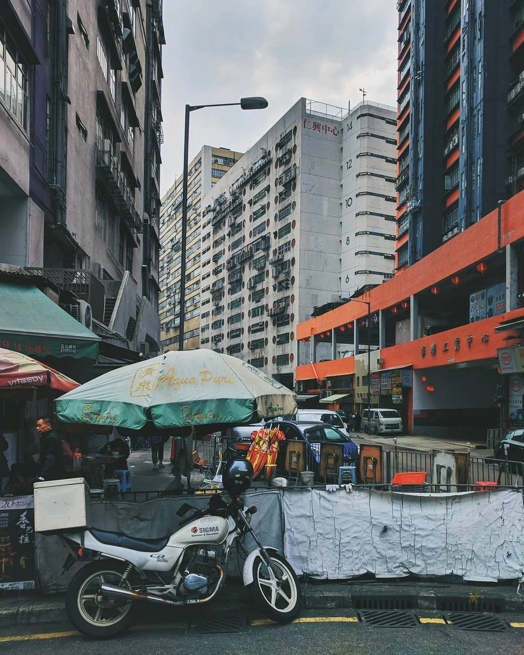 Street scene in an urban area with high-rise buildings, a parked motorcycle, and a large umbrella covering street vendors and small shops.