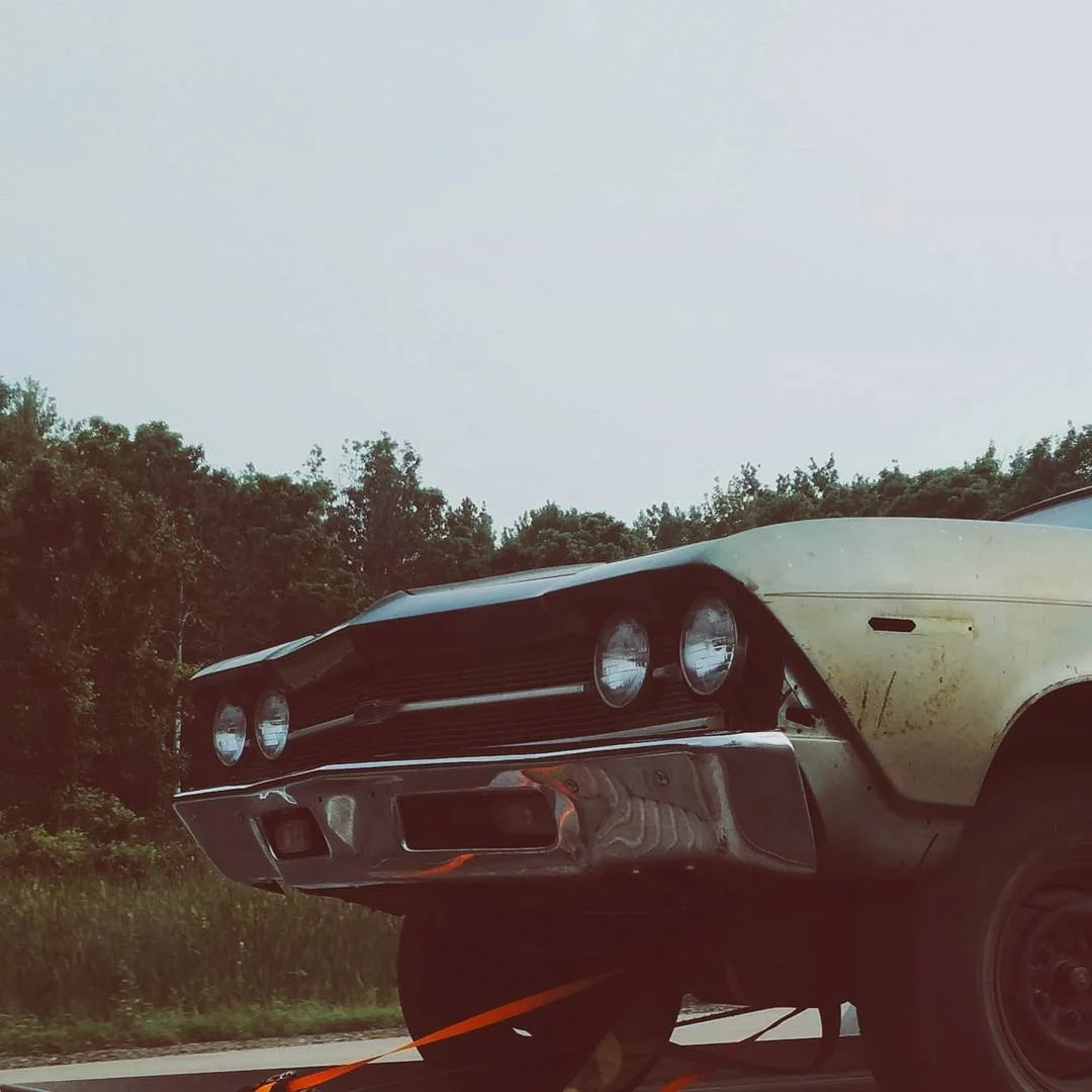 Close-up of a vintage yellow and black car with a damaged bumper, driving on a road with trees in the background.