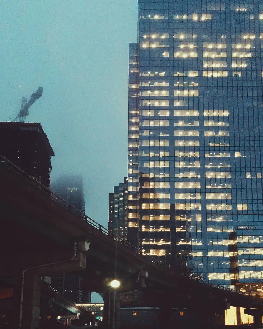 High-rise glass office building with illuminated windows at dusk, city street with elevated train tracks in foreground.