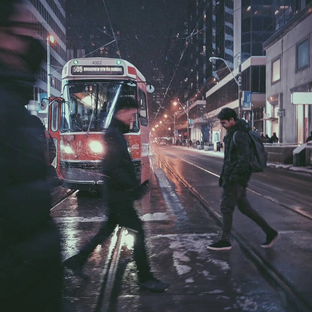 People crossing wet city street at night near a parked streetcar with overhead wires, surrounded by tall buildings.