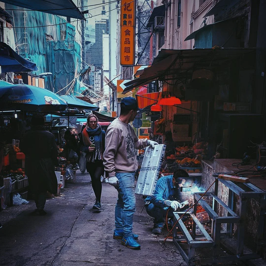 A narrow street market with vendors and shoppers, some behind umbrellas, with signs in Asian characters, and a worker welding metal near a fruit stand.