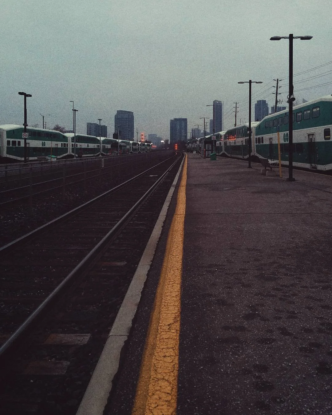 City train station with two train tracks and parked double-decker commuter trains on either side, with city skyscrapers in the background at dusk.