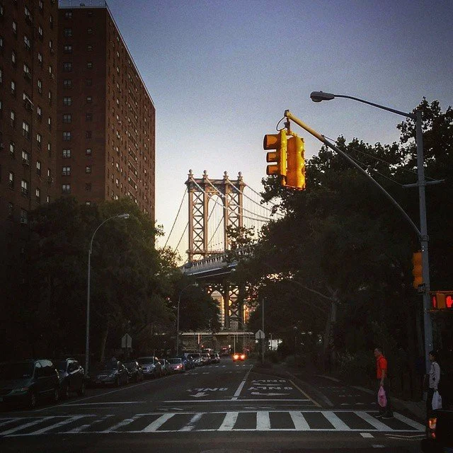City street with traffic lights, cars parked along the side, trees, and a view of the Manhattan Bridge in the background during sunset.