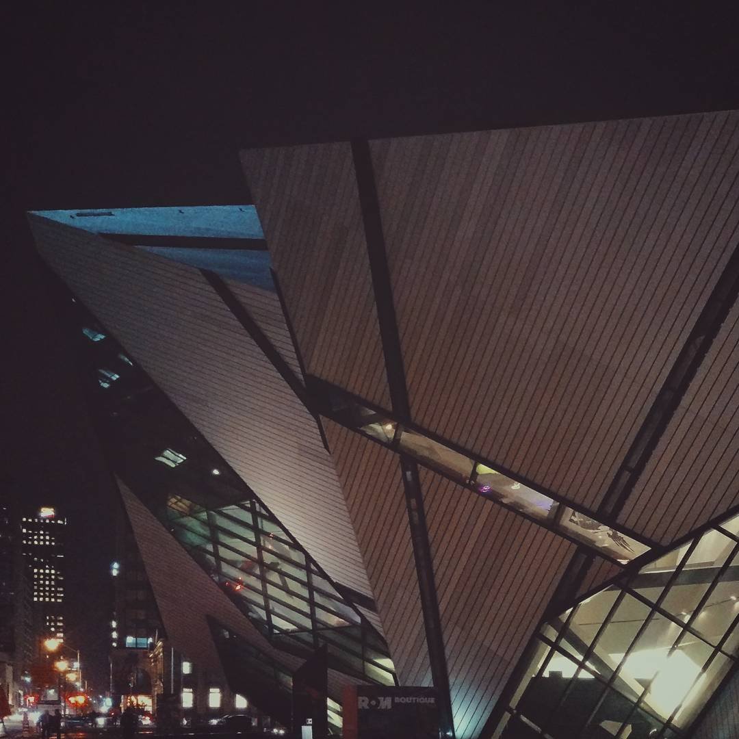 Night view of a modern building with angular, slanted wooden and glass panels, illuminated from inside, with city lights in the background.