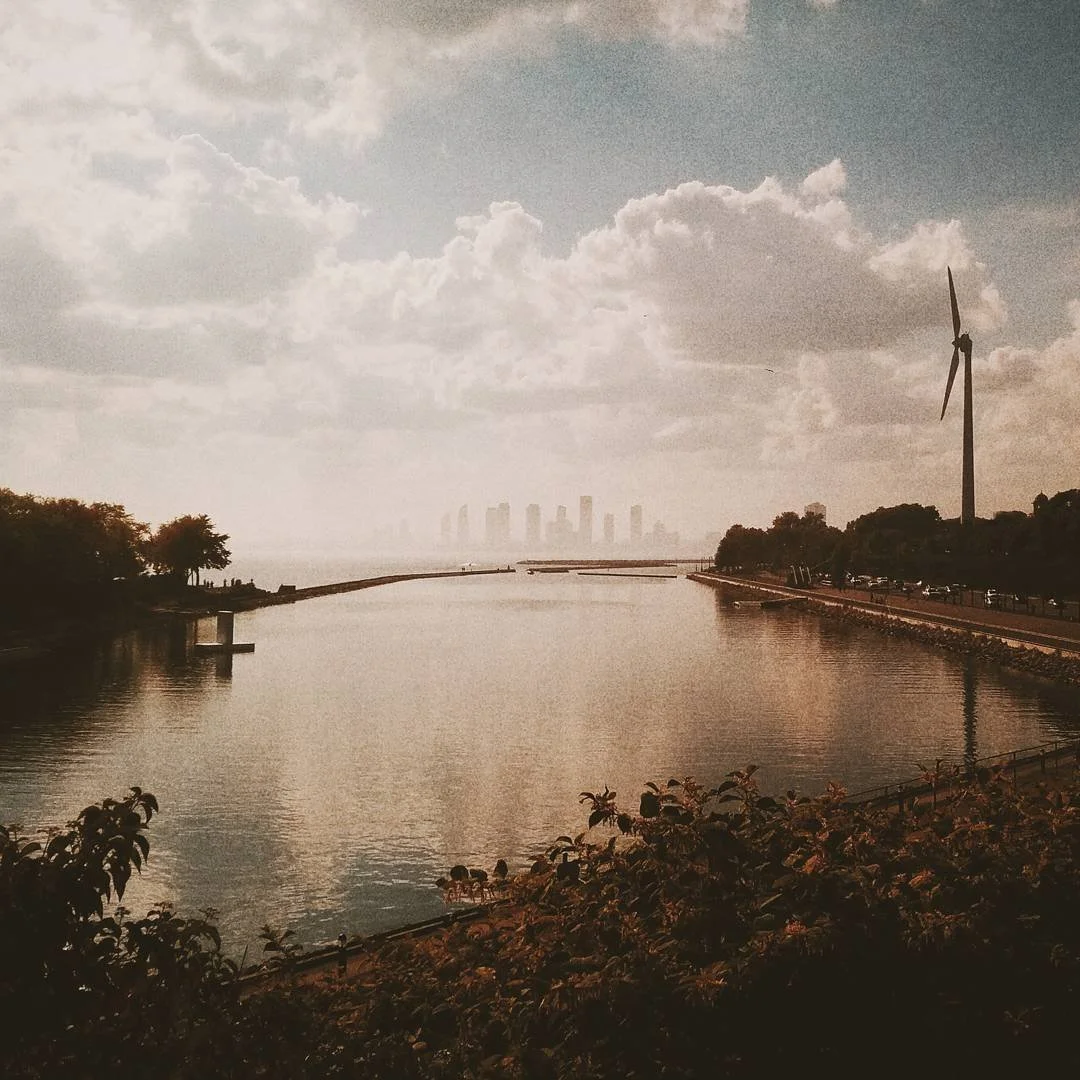 A calm river scene with a cloudy sky, wind turbines, and a city skyline in the distance.