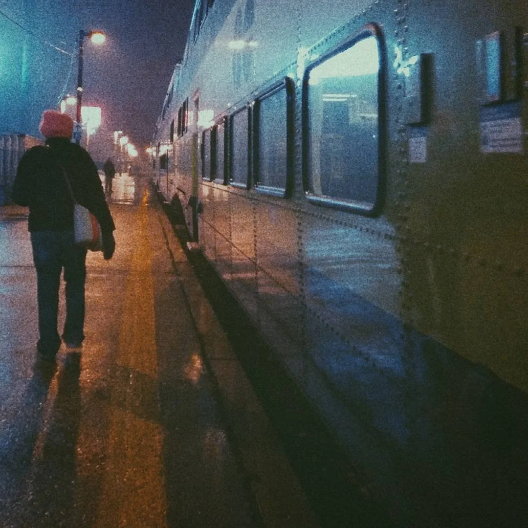 A person walking along a wet city street at night, with a large bus parked on the side and streetlights illuminating the scene.
