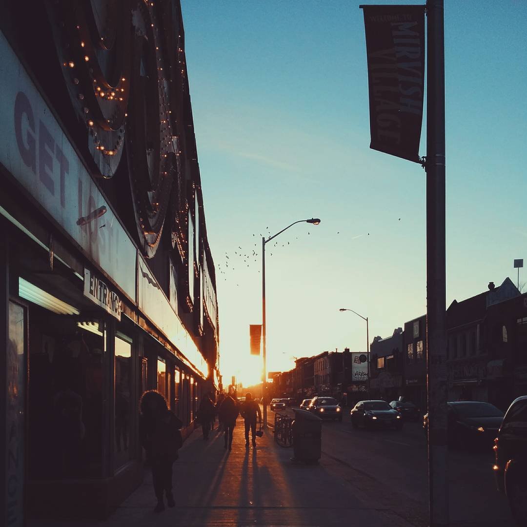 City street at sunset with pedestrians walking and cars parked along the side, building facades and streetlights visible, sky clear with a few birds flying, and a sign for Miry’s List.