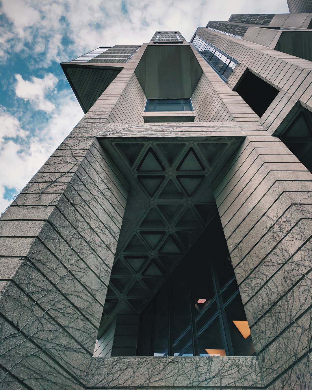 Low-angle view of a modern high-rise building with geometric design features, glass windows, and concrete walls against a partly cloudy sky.