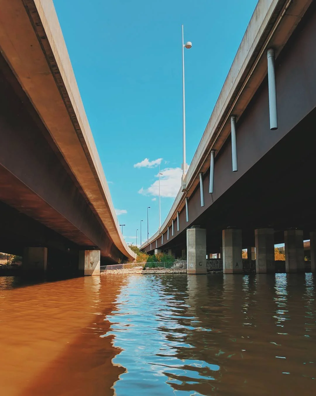 View of two parallel bridges over a body of water, under a bright blue sky with some clouds, showing the underside of the bridges and streetlights.