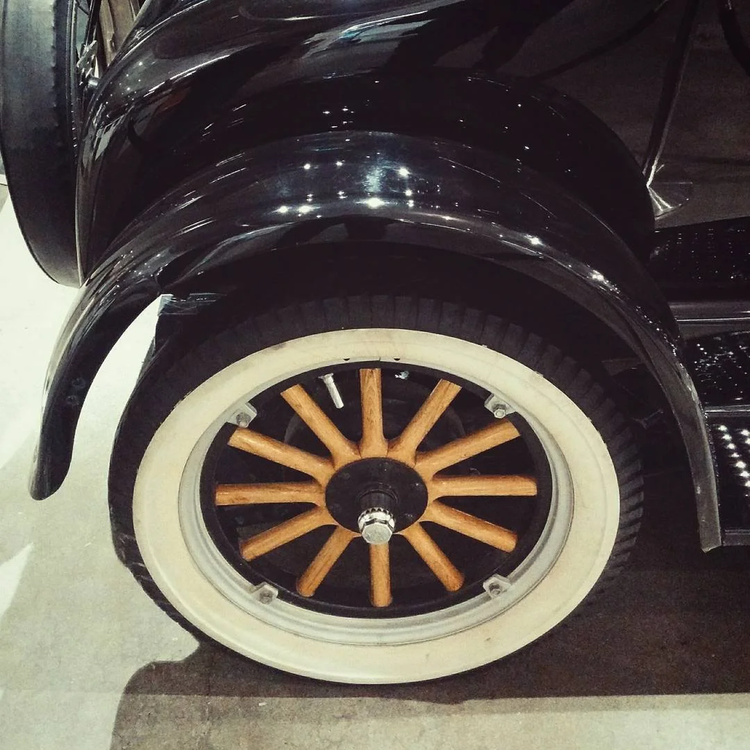 Close-up of a vintage car wheel with a wooden spoke design, a white tire, and a black fender.