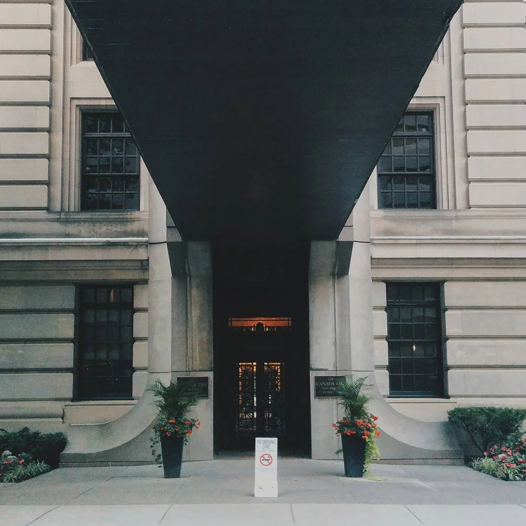 Front entrance of a large stone building with a dark awning overhead, potted plants with flowers on either side, and a no smoking sign on the sidewalk.