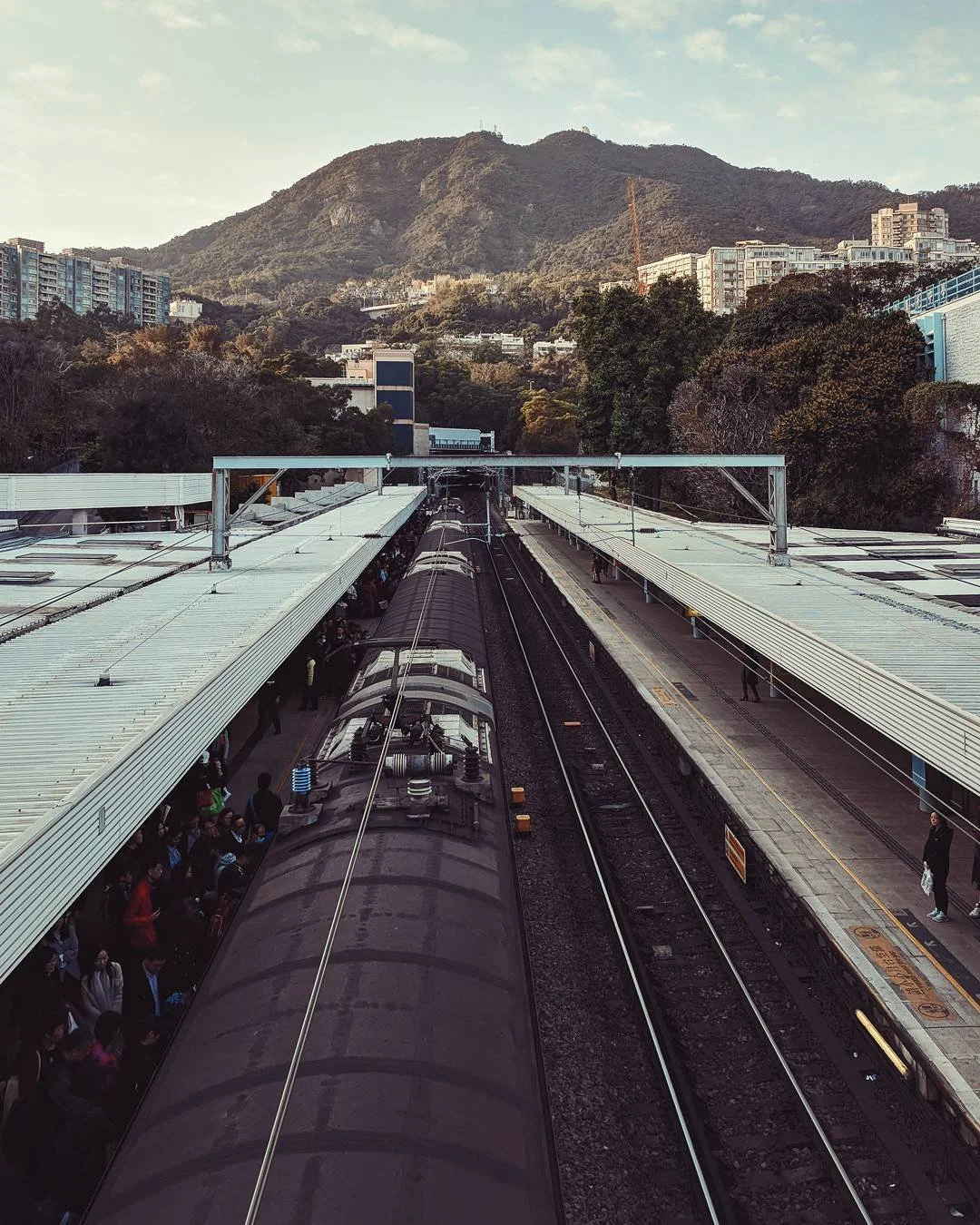 View of a train station with platforms on both sides and a train in the middle, surrounded by trees and mountains in the background on a partly cloudy day.