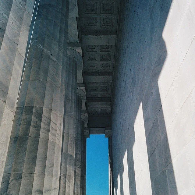 Looking up at the Lincoln Memorial in Washington, D.C., showing the tall stone columns and the roof with patterned panels, with a view of the blue sky through the opening at the top.