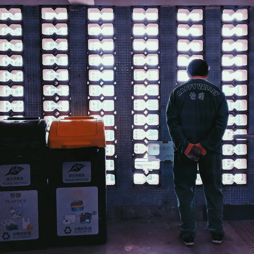 A man stands with his back facing a patterned wall with bright light coming through, wearing a jacket with 'HAPPY VALLEY' written on the back, and is separated from recycling bins labeled for plastics.