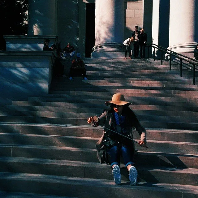 A woman wearing a large hat, sunglasses, and dark clothing sitting on steps outside a building, looking at her phone, with others in the background near large columns.