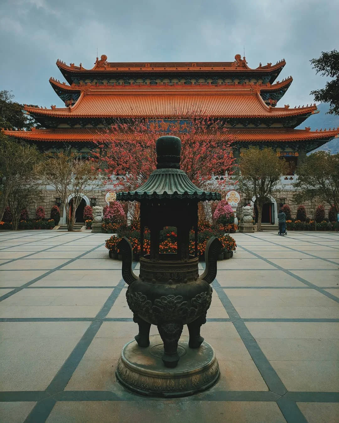 Traditional Asian temple with red pagoda-style roofs, adorned with trees and flowers in front, and a large incense burner in the foreground.