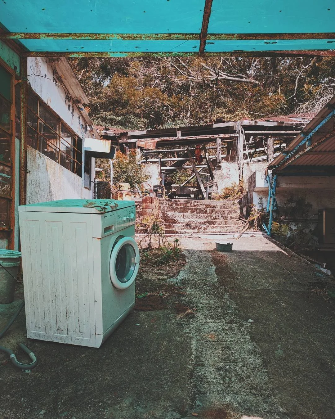An outdoor area with an old washing machine on a concrete ground, surrounded by overgrown plants and rusted structures, with stairs leading up to a higher level and trees in the background.