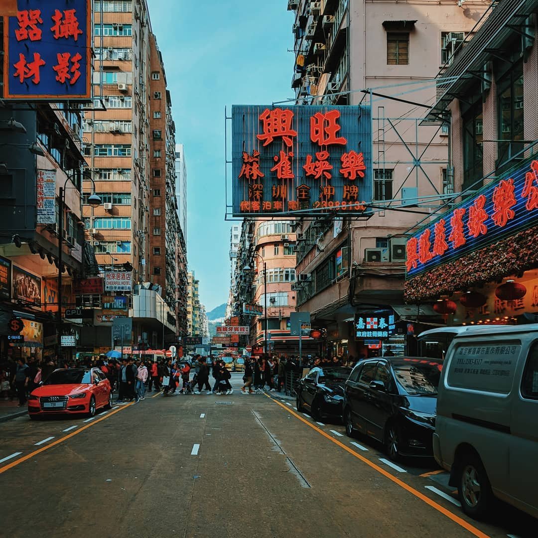 Street scene in an Asian city with tall buildings, neon signs with Chinese characters, and pedestrians crossing the street. Cars are parked along the sides.