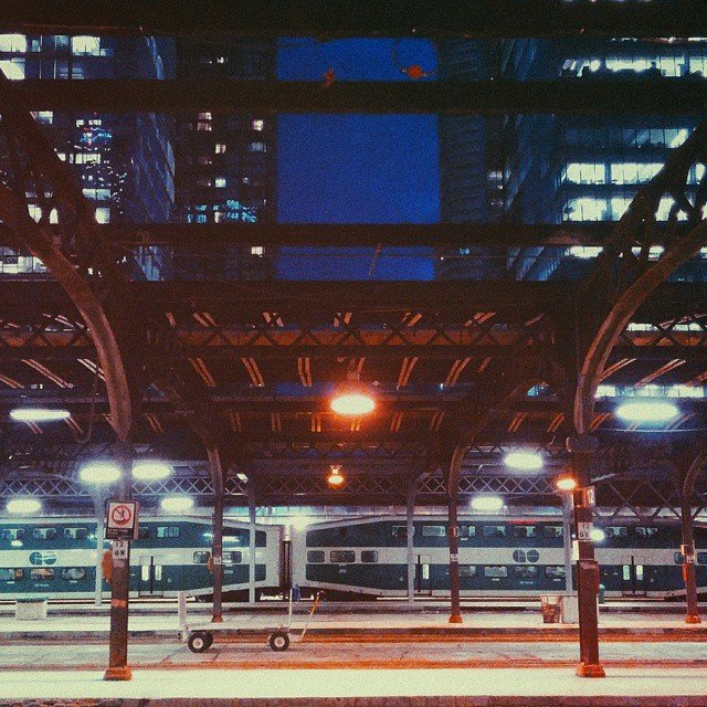 Empty train station platform in an urban setting at night, with a train in the background and city lights overhead.