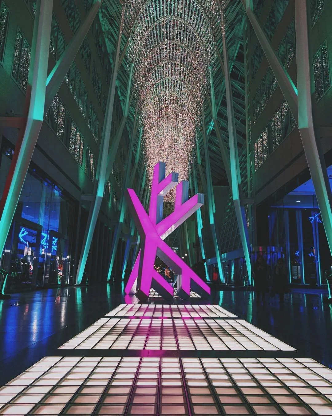 Colorful modern art installation inside a building with tall glass walls. The installation features illuminated pink and purple geometric shapes on a grid walkway, with ceiling lights creating a glowing arch pattern.