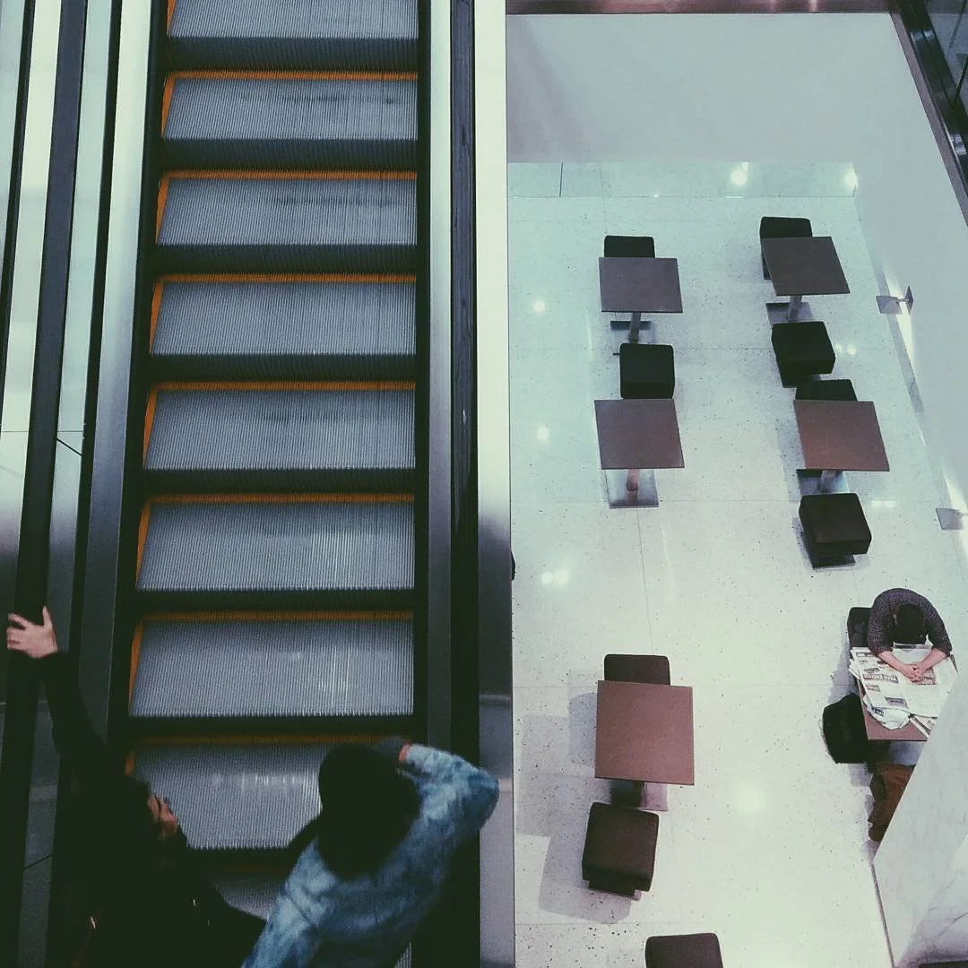 An aerial view of an escalator on the left side with two people riding it, and a seating area with chairs and tables on the right side with one person sitting at a table reading newspapers.