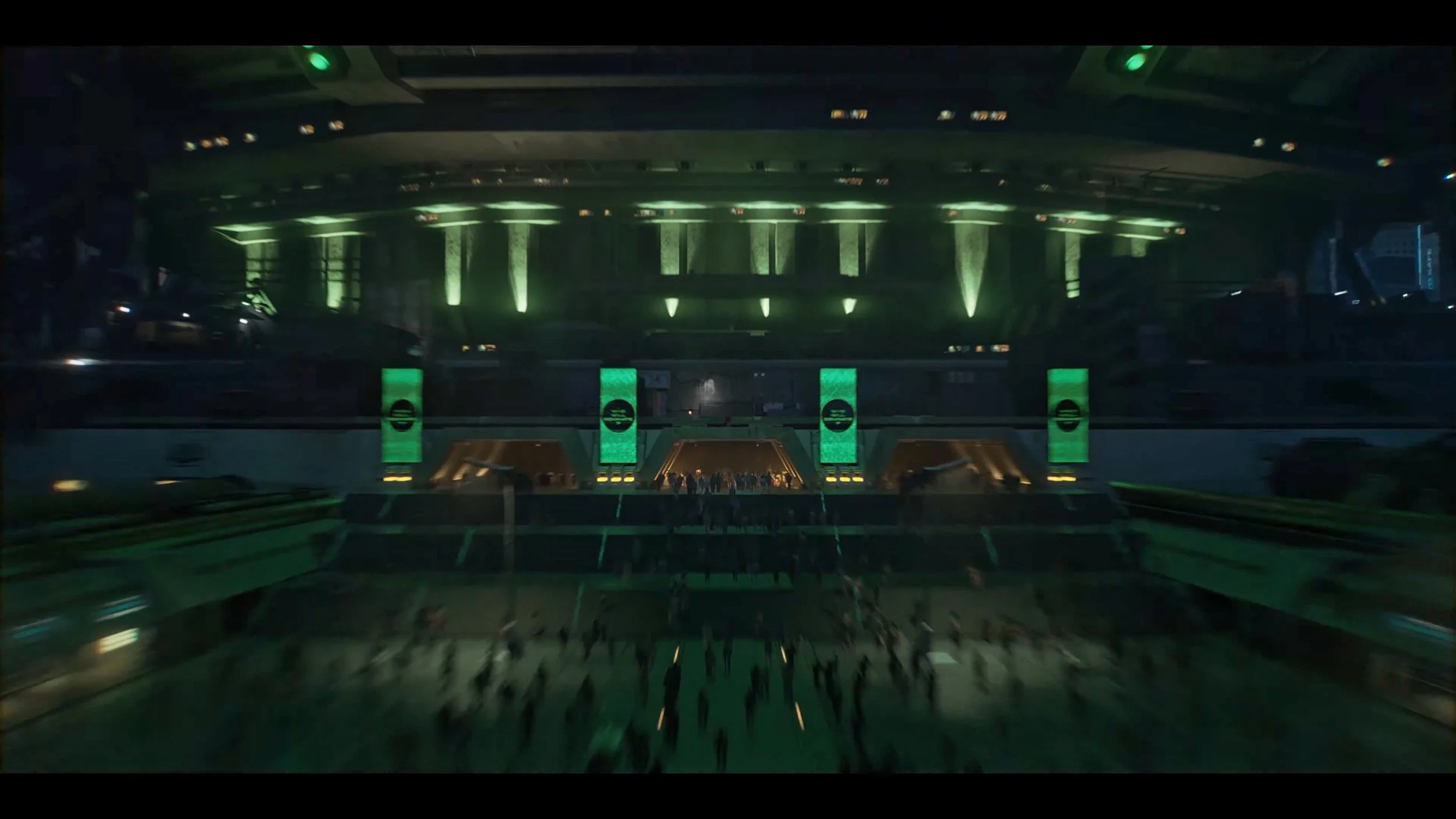 A dark, futuristic interior of a large building or spaceship with green lighting. People are gathered near the center, walking or standing, with stages or platforms on either side and multiple levels, creating a sci-fi or cyberpunk atmosphere.