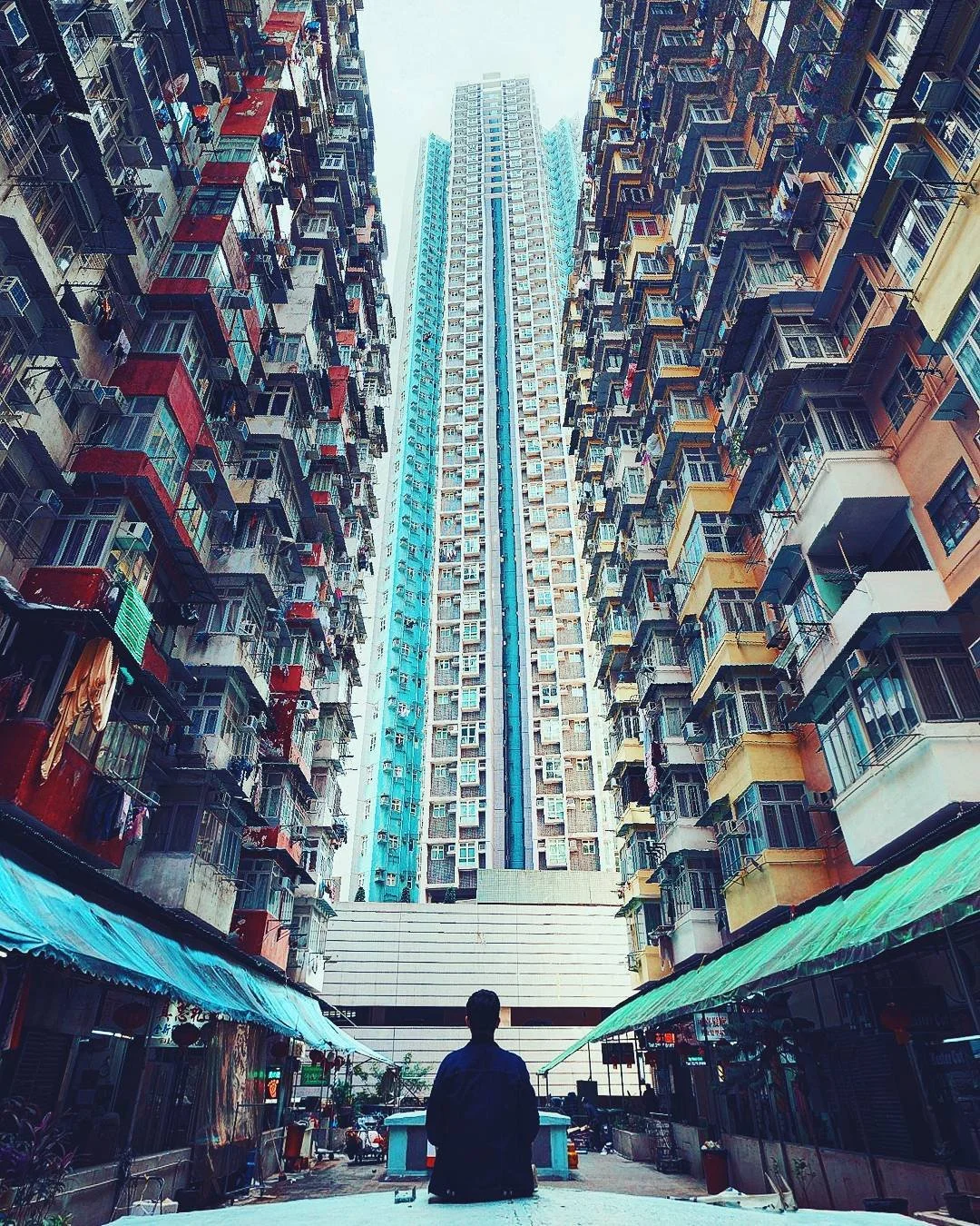 A person sitting in an urban alleyway surrounded by tall apartment buildings with laundry hanging outside their windows.