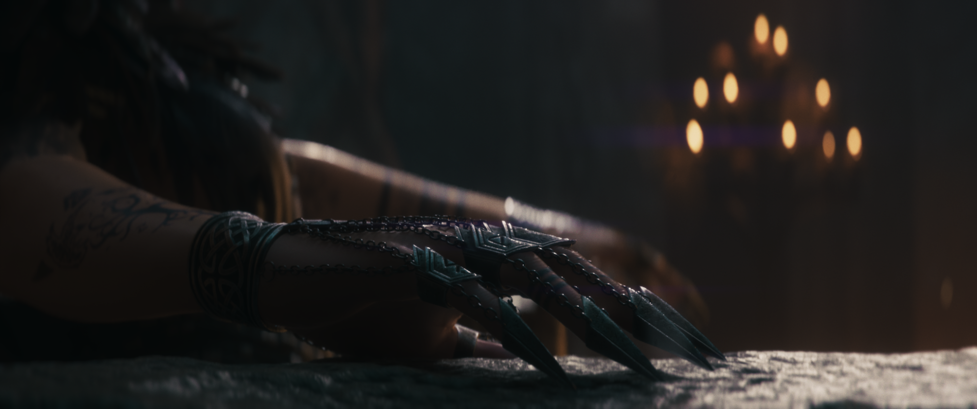 Close-up of a hand with long, sharp metallic claws and tattoos on the arm, resting on a surface in a dark room with blurred warm lights in the background.