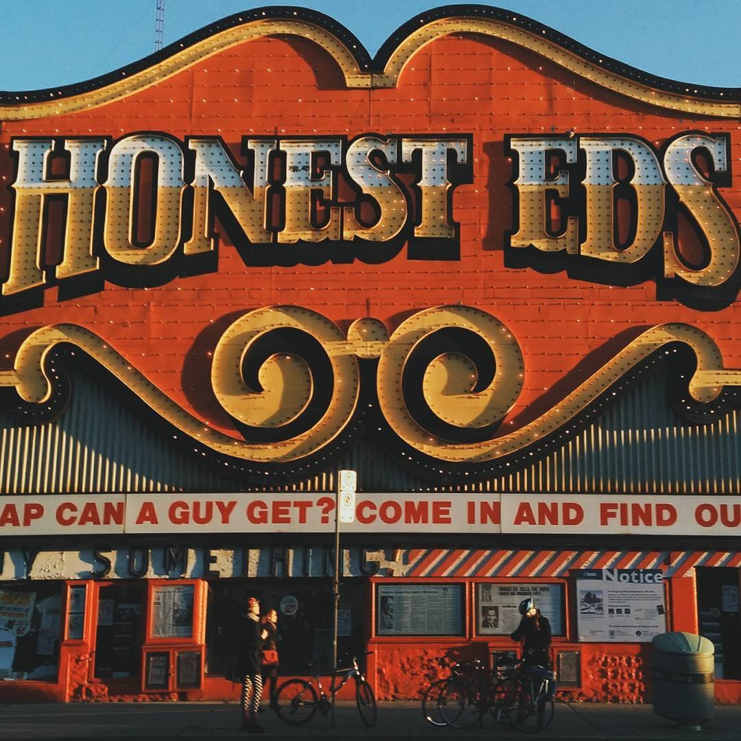 The front of a red building with large illuminated sign spelling 'HONES E' and part of the word 'STEPS' visible, with people and bicycles in front, and a blue sky overhead.