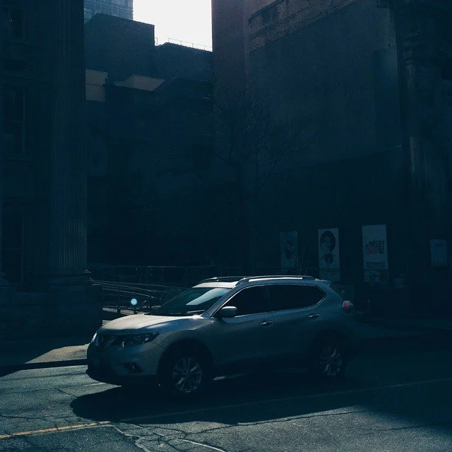 A silver SUV parked in a city lot with a steep building wall and posters on the wall in the background.