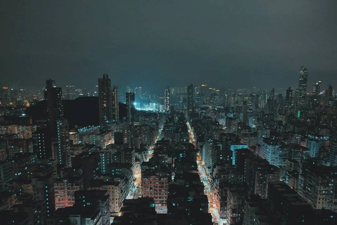 Night view of a cityscape with tall buildings, illuminated streets, and a dark cloudy sky.