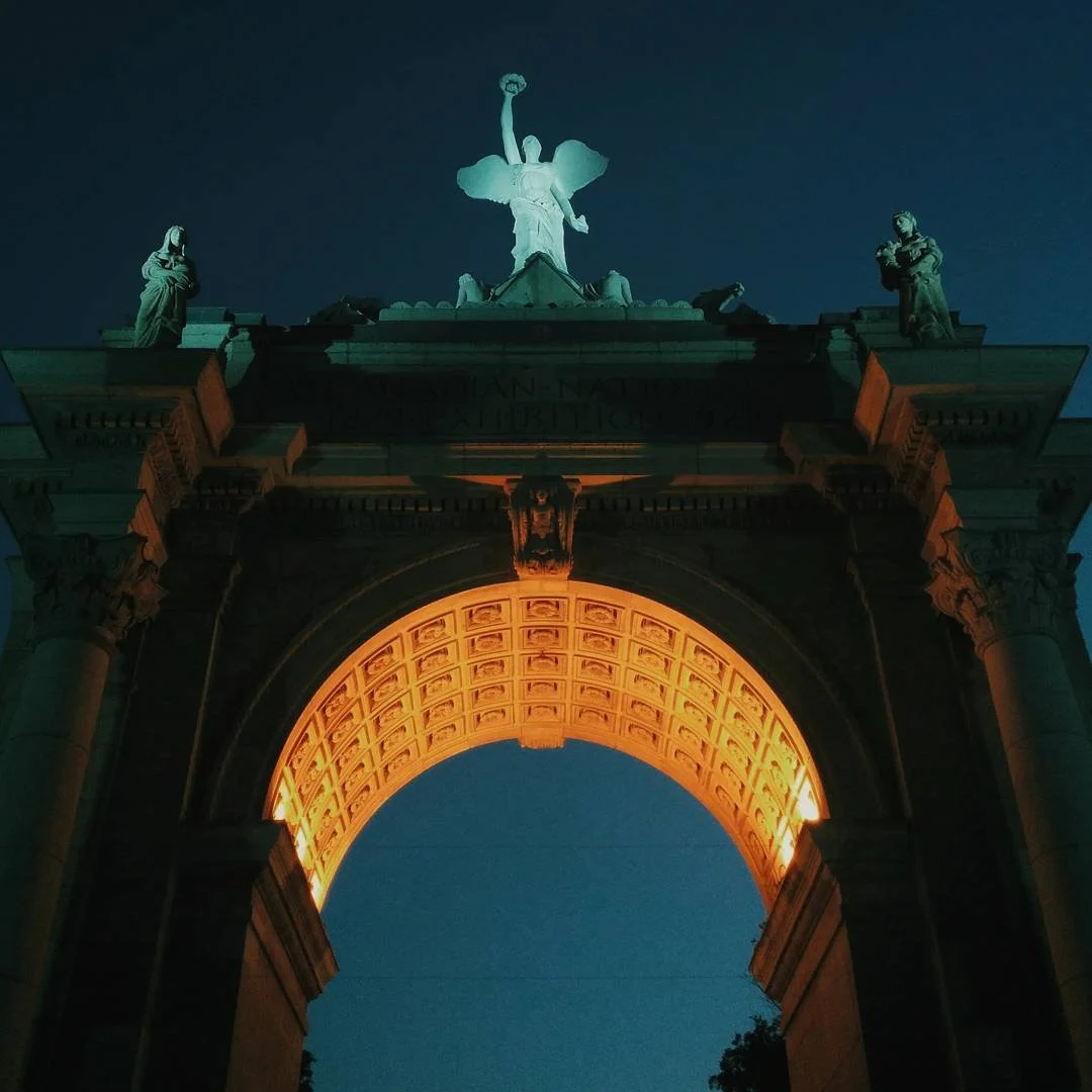 An illuminated archway with sculptures on top, including a central figure holding a torch, at night.