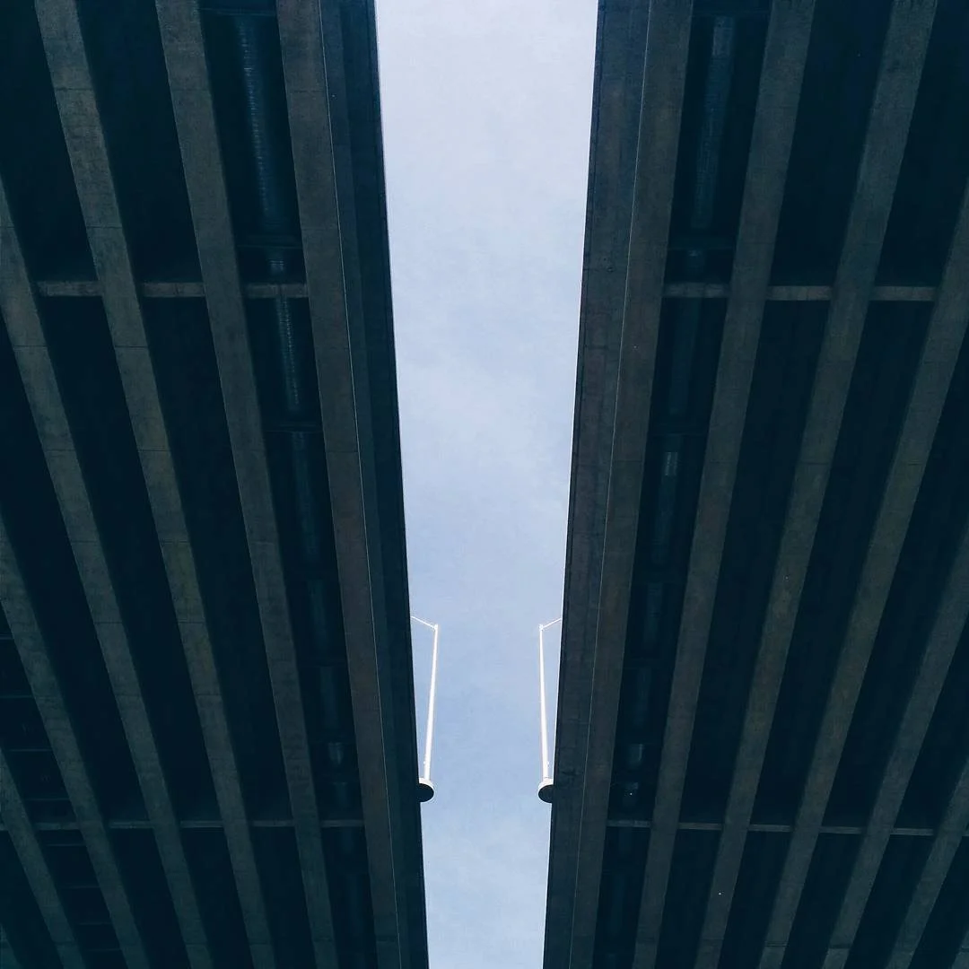 Looking up between two dark modern buildings with metallic panels and beams, showing the blue sky and a thin strip of clouds above.