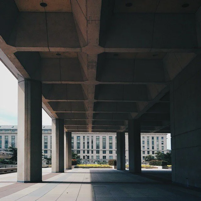 Underneath a large concrete structure with multiple supporting columns and geometrically patterned ceiling, with a view of a modern office building and sidewalk in the background.