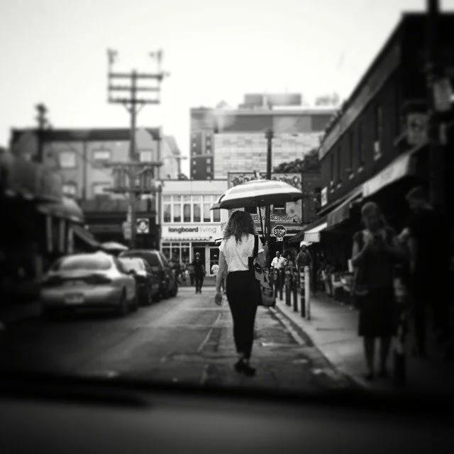 A woman walking down a city street with an umbrella, surrounded by cars and pedestrians, with buildings and a train in the background, in black and white.