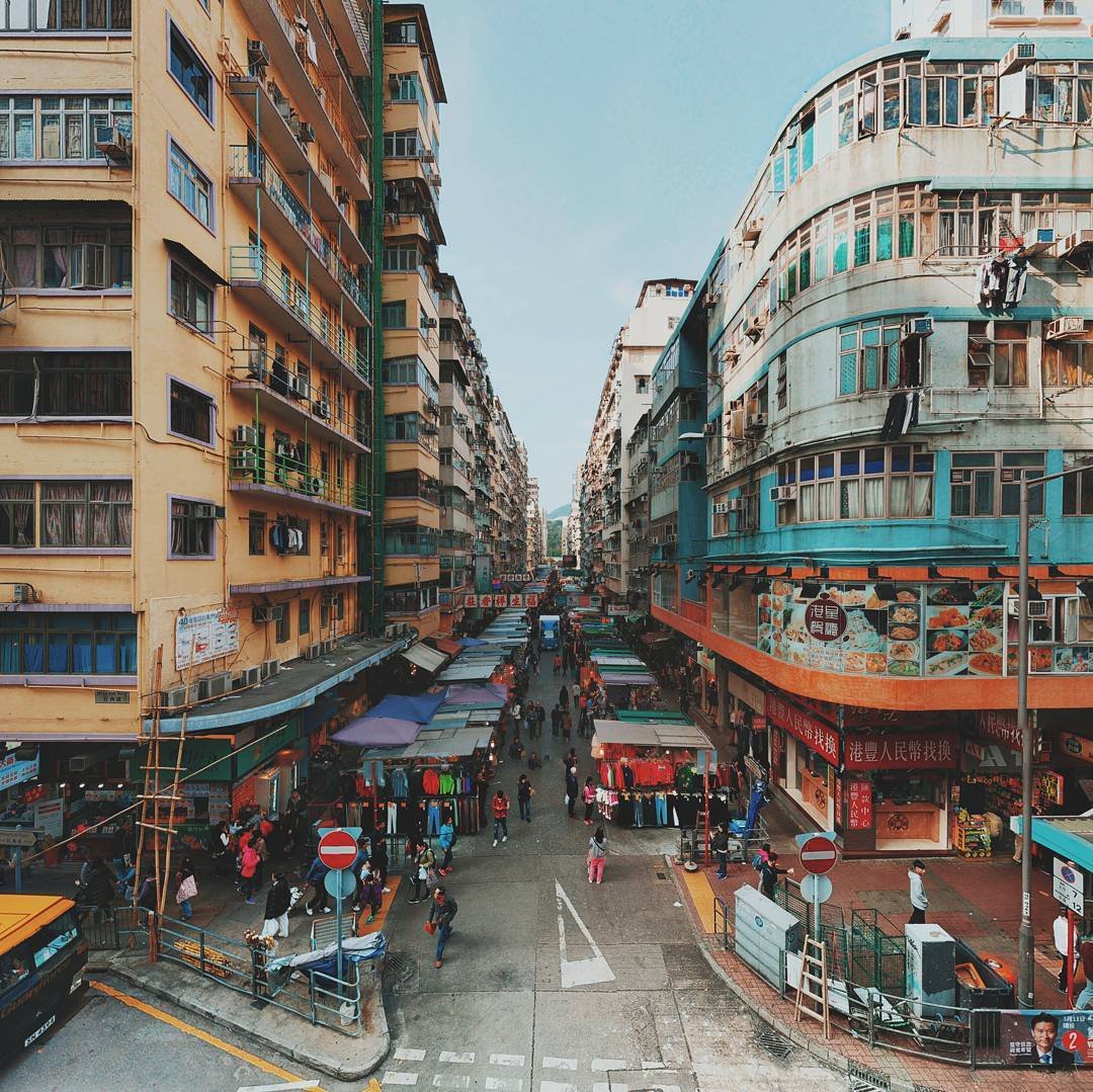 A busy street market scene in an urban area with high-rise buildings on both sides, storefronts with colorful signage, and outdoor stalls selling clothes and food. Pedestrians walk along the street, some browsing stalls and others crossing at the intersections.