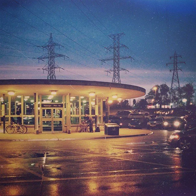 A parking lot outside a transportation hub at night, with a round glass building entrance and multiple parked cars. Overhead, there are power lines and electrical towers under a starry sky.