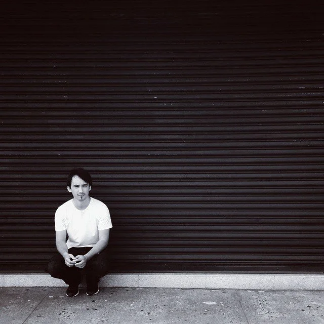 A young man with dark hair, wearing a white t-shirt and dark pants, sitting on the sidewalk in front of closed, horizontal metal roller shutters, looking at the camera.