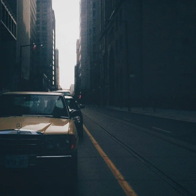 Old classic yellow Mercedes-Benz car parked on a city street with tram tracks, tall buildings on both sides, and a distant overcast sky.
