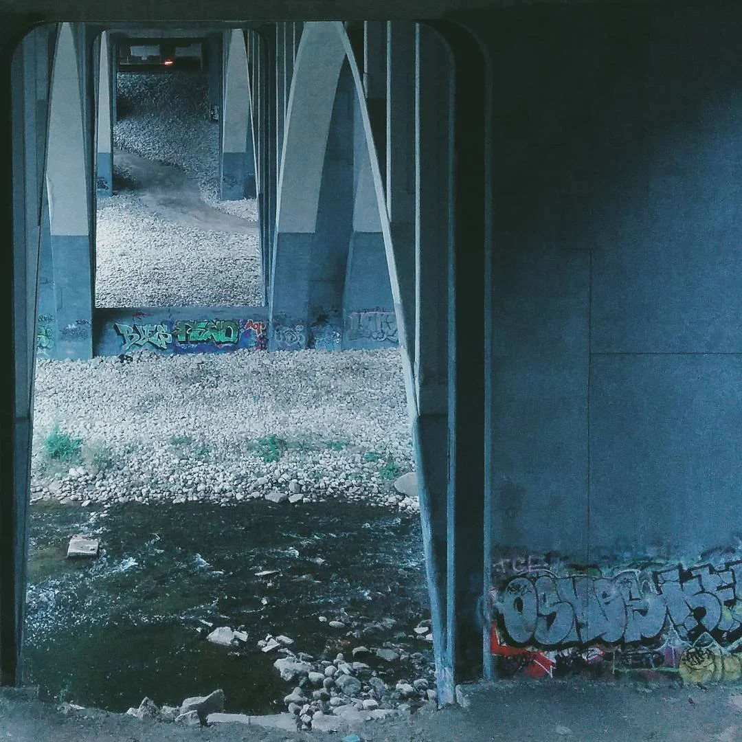 View of the underside of a bridge with graffiti, showing a stream of water and gravel below.