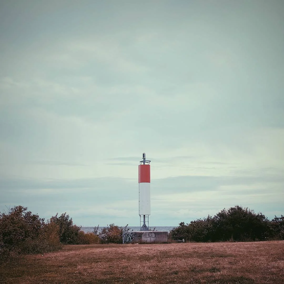 An open field with grass and bushes, and a tall red and white lighthouse in the distance under a cloudy sky.